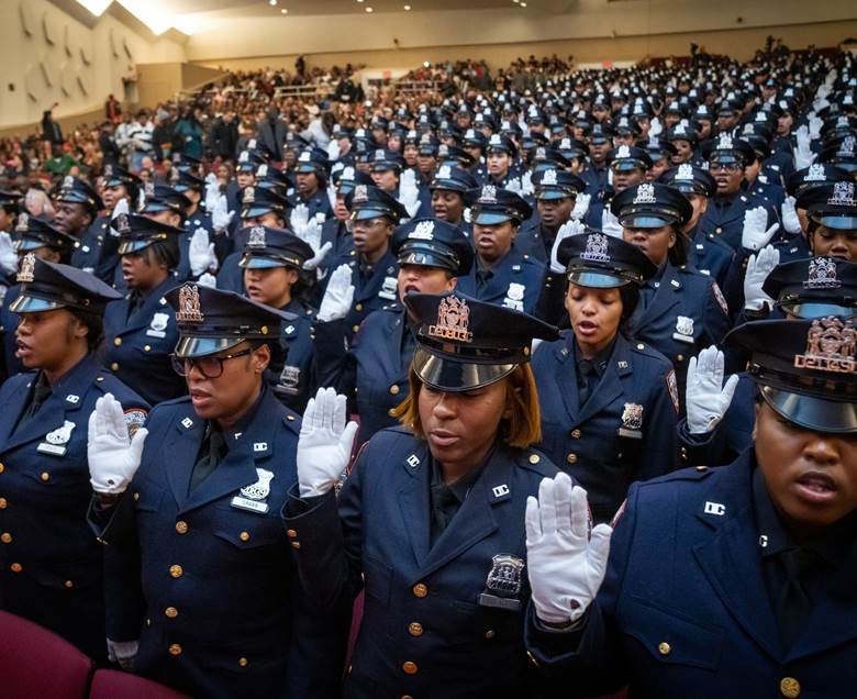 A large group of people standing in a row, all wearing blue uniforms and holding their hands up. Description generated by AI