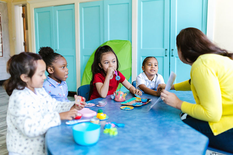 Photo of children and their teacher sitting around a table with toy tea party playset.