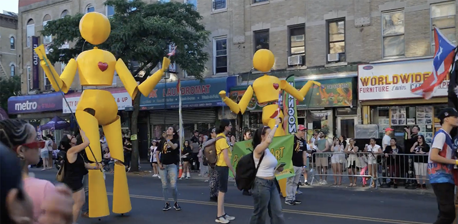 Community parade featuring two bright yellow Sunny puppets moving along a city block as people march and gather nearby.