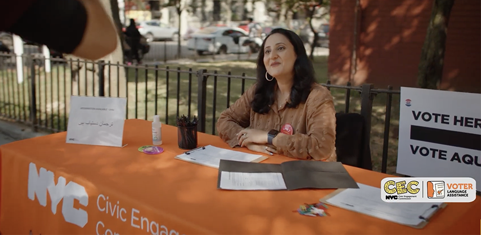 Vote in Your Language! PSA - Civic Engagement Commission - Woman seated at a Civic Engagement Commission voter assistance table outdoors, smiling toward a visitor beside a sign that says &ldquo;Vote Here / Vote Aqu&iacute;.&rdquo;