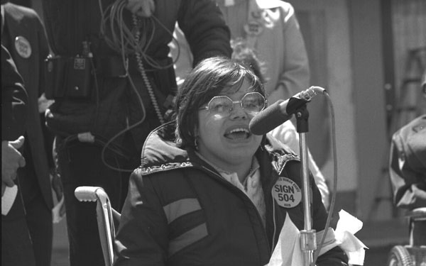 Black-and-white photo of Judith Heumann, a disability rights activist, speaking into a microphone at a public protest. She is seated in a wheelchair, wearing glasses, and a crowd is visible in the background."