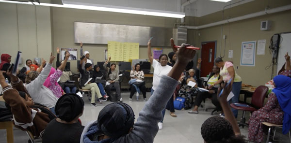 a workshop part of the Mothers Matter program. A woman is standing up leading the workshop with her hand in the air, and the participants are sitting around her in a circle, most of them also raising their hands.