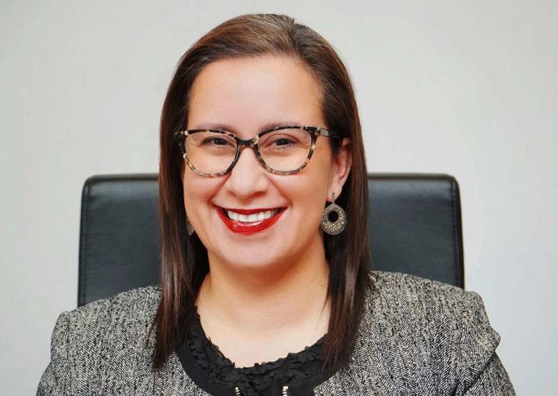 This is a headshot of Dr. Helen Arteaga Landaverde sitting and smiling warmly at the camera. She is wearing glasses, has straight, shoulder-length hair, and she is dressed in a grey and black outfit.

