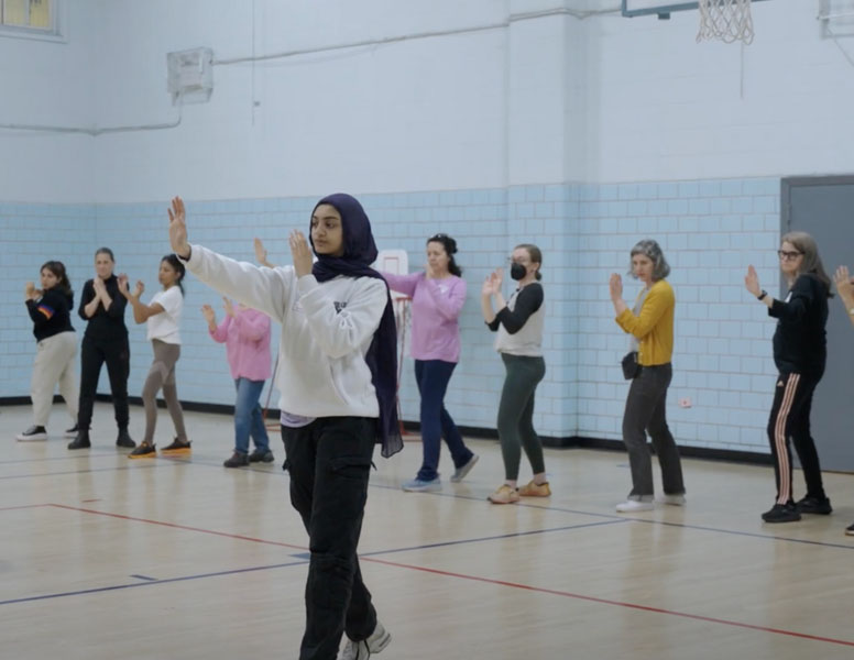 To	The photo shows a self-defense workshop led by the organization Malikah. It features a young woman showing a self-defense move to a line of women behind her in a gym.