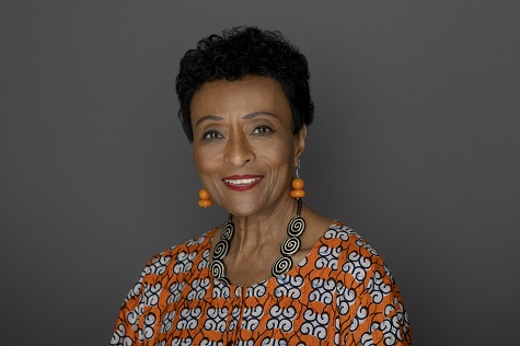 Headshot photo of Peggy Shepard smiling at the camera with a patterned orange top, a necklace, and orange earrings.