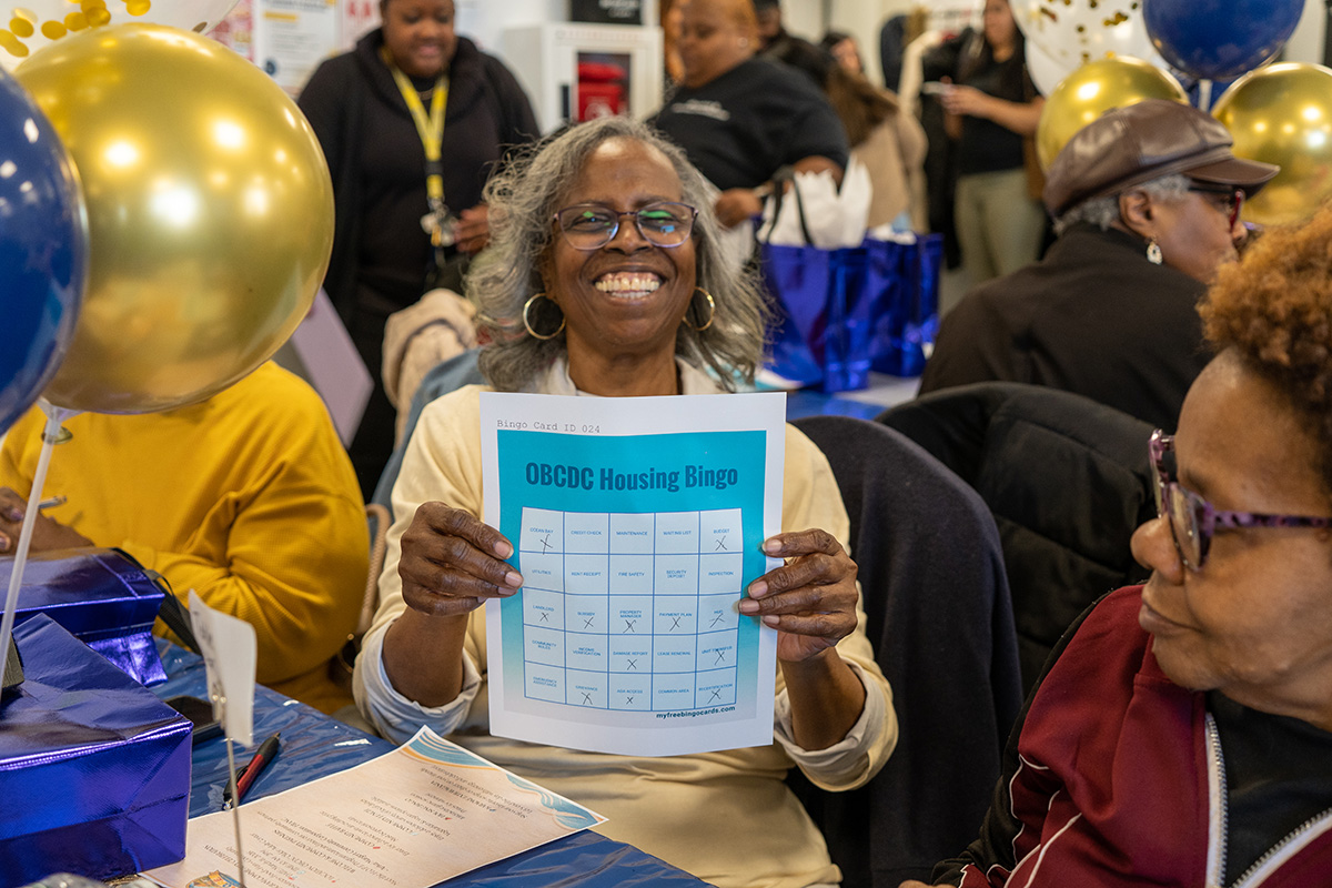 Photo of an older adult woman smiling holding up a housing-themed bingo card, sitting at a table with blue and gold balloons and other older adults.
