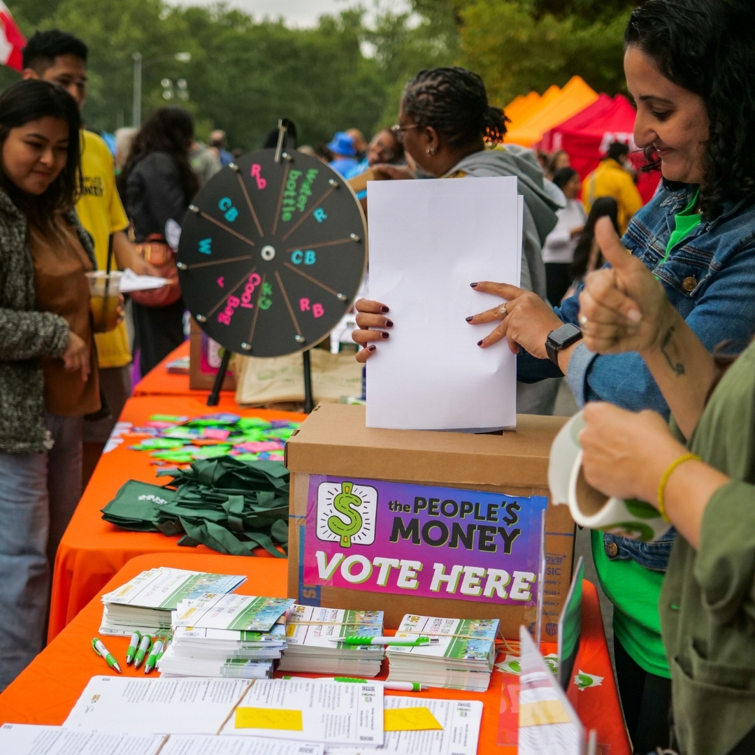 Photo of people standing at tables outside that have orange tablecloths and ballots, People's Money merch, a spinning wheel game, and a ballot box on top. A woman is smiling as she puts ballots into the ballot box.
