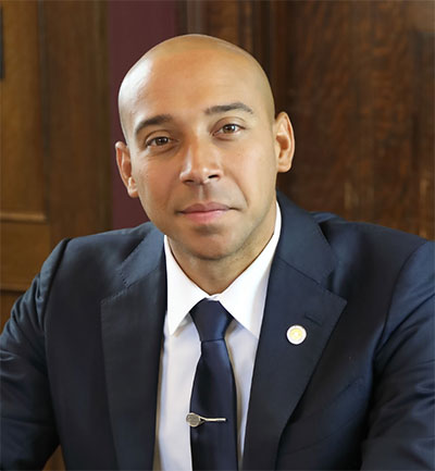 Headshot photo of Alister Martin smiling and wearing a navy blue suit and tie in front of a brown background