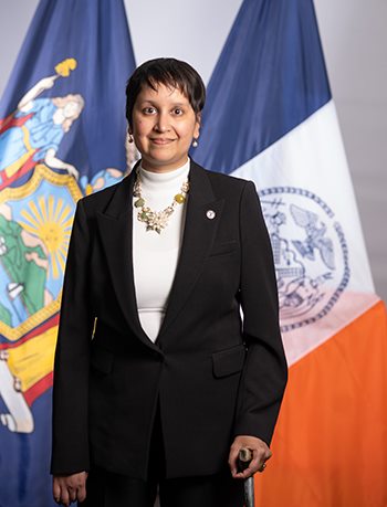 Photo of Commissioner Nisha Agarwal in a black suit with a white shirt and necklace, standing in front of the NYC flags.