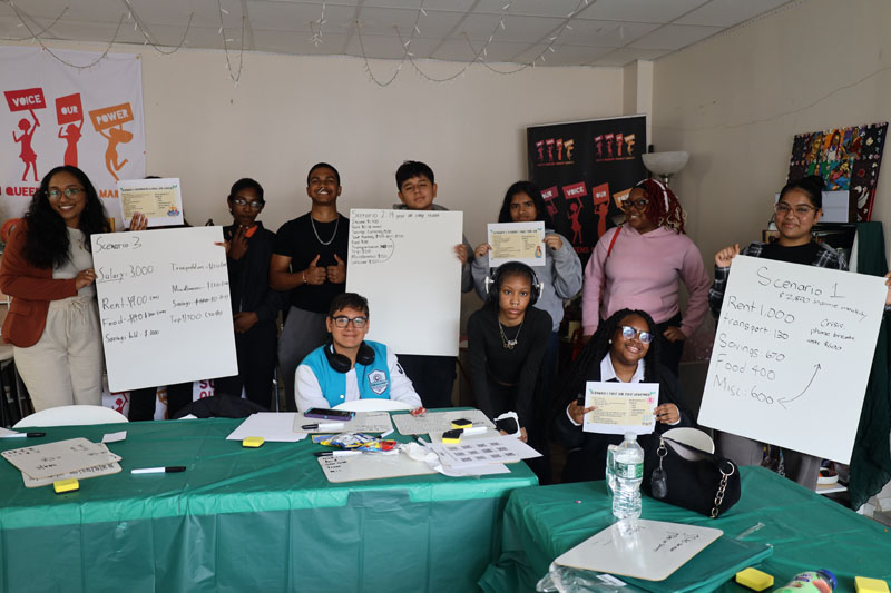 Photo of approximately 10 youth project participants holding up large pieces of paper with a budget activity on it as they sit and stand around tables with green tablecloths. 