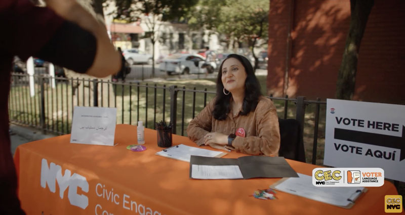 Thumbnail image for Vote in Your Language PSA video, with a woman sitting at a table with an orange tablecloth that says “NYC Civic Engagement Commission” as she greets someone coming up to the table. There is a sign that says “Vote here / Vote aqui
