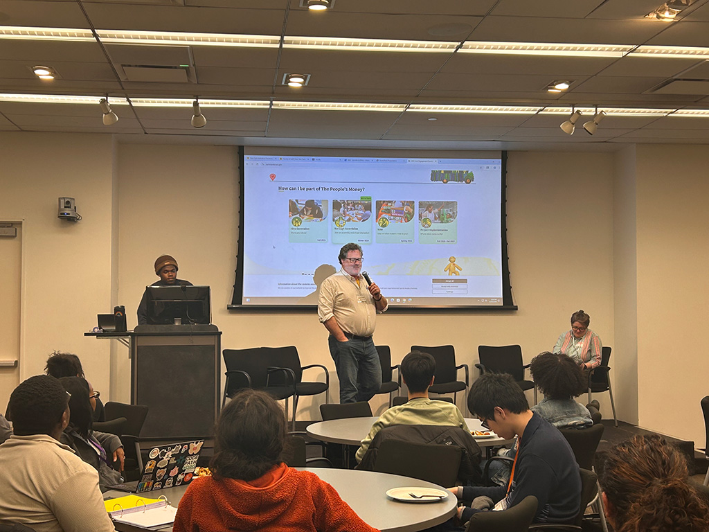 This photo shows a CEC staff member holding a  microphone presenting to a group sitting at tables during the Datathon mixer. There is a large screen at the front of the room showing the homepage of the CEC's Participate website.