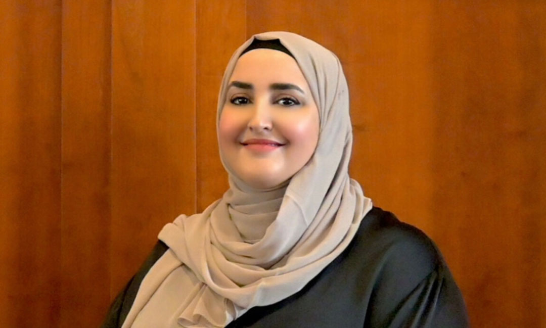 This is a headshot photo of Somia El-Rowmeim wearing a beige hijab and black shirt in front of a brown background..