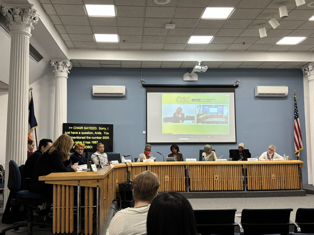 Photo from the CEC Commissioners' meeting, showing commissioners and a CEC staff member sitting around a panel on stage at Spector Hall as the CEC staff member presents. There is a projector screen behind them showing the presentation, as well as a screen showing closed captions.