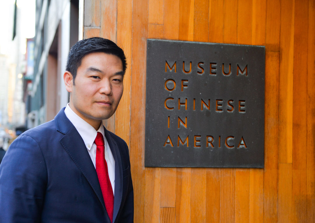 o	Photo of Michael Lee in a blue suit and red tie posing in front of a Museum of Chinese in America sign.