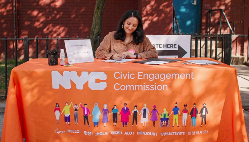 Person sits at orange table with NYC Civic Engagement Commission banner in various languages waiting to assist voters with interpretation services