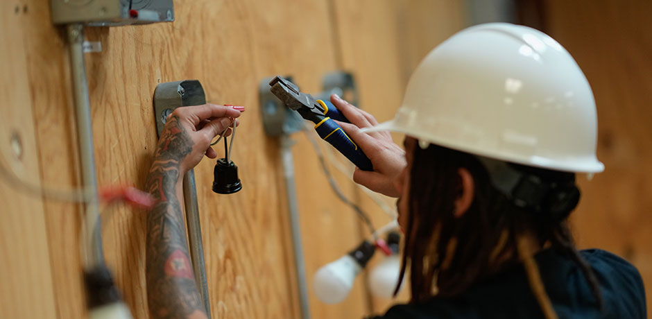 A male construction apprentice wearing a white hardhat practices a task related to installing an electrical outlet in a classroom
                                           