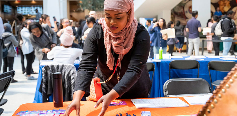A woman wearing a hijab sets up her table with flyers and pens during a job fair that is full of attendees
                                           