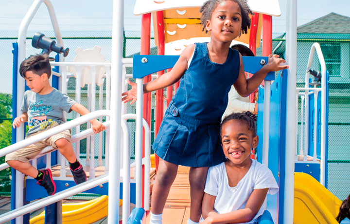two girls pose on a playground while boy slides down a post in the background
                                           