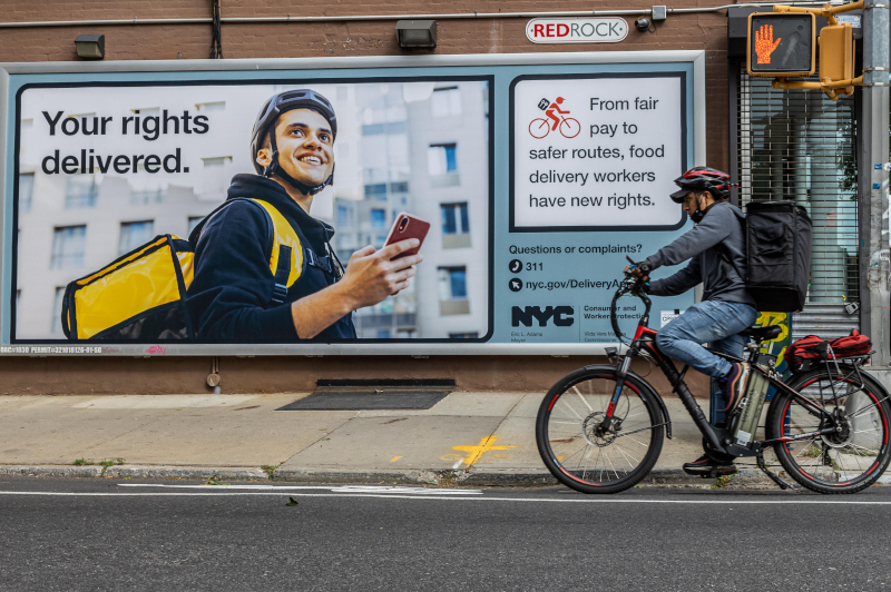 Food delivery worker on bicycle rides pass large billboard about worker rights
                                           