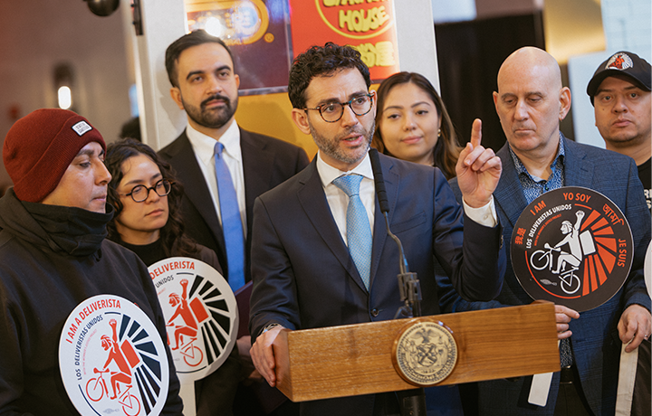 DCWP Commissioner Sam Levine speaks at a podium along with Mayor Zohran Mamdani, councilmember Harvey Epstein, and members of Los Deliveristas Unidos
                                           