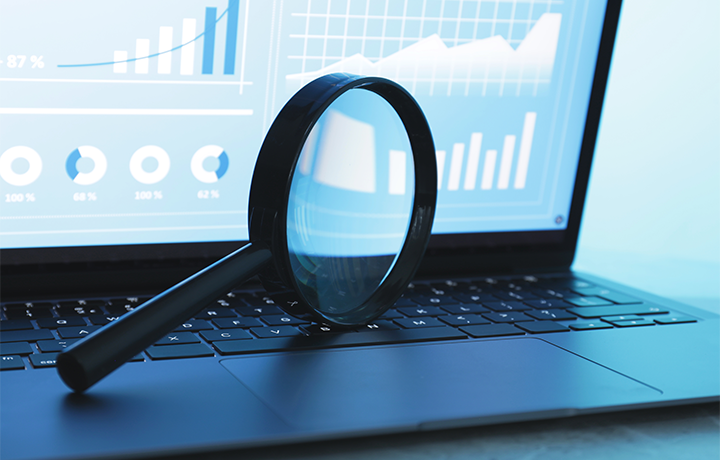 Magnifying glass sits in front of a computer screen showing analytics and graphs
                                           