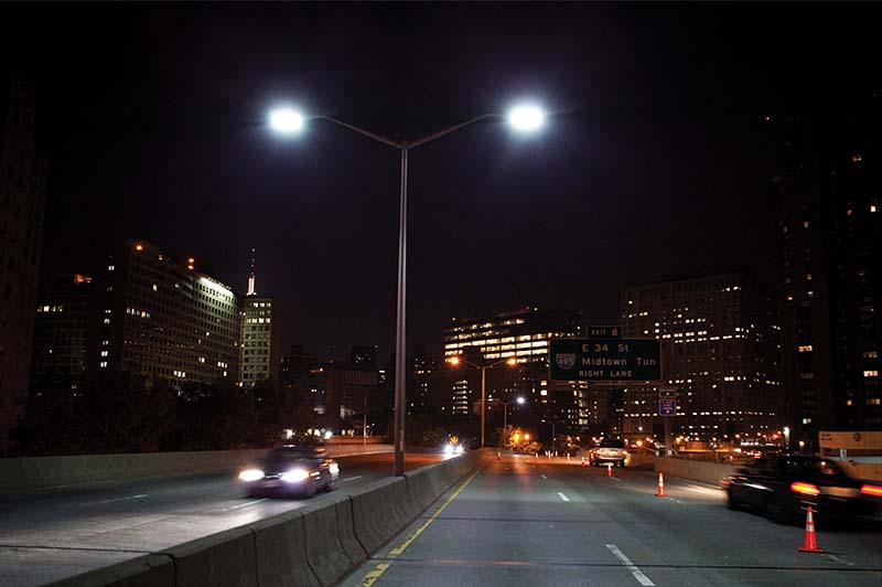 Six lane highway at night illuminated by street lights and the light from cars on the highway. The City is in the background