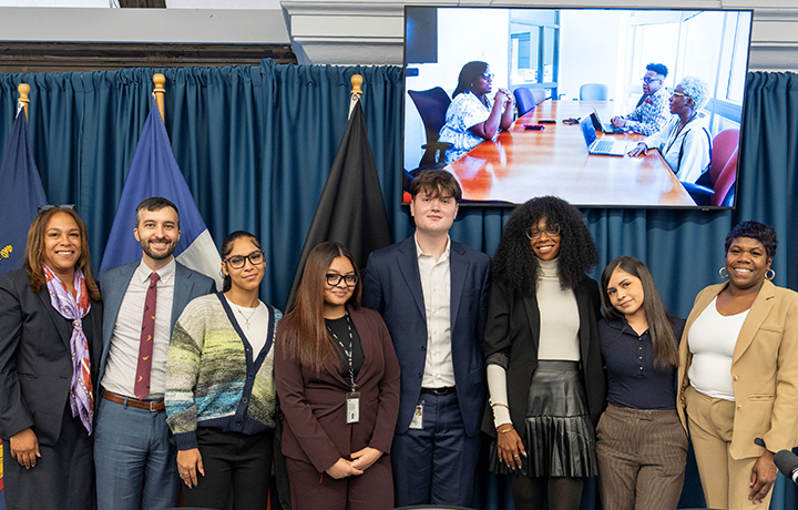 Youth Apprentices and city officials gather in front of several flags and a television monitor and a blue curtain for a panel discussion.
                                           