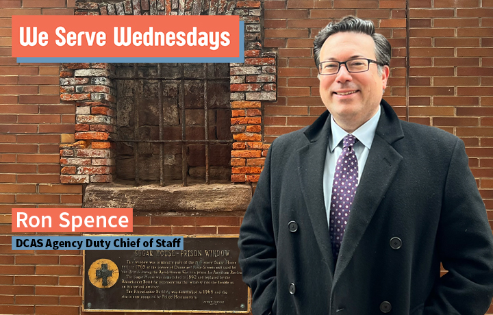 Smiling man in a suit and tie standing in front of a brick wall.
                                           