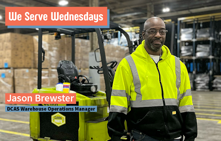 Man in a reflective vest, by a forklift in a warehouse with boxes behind him.
                                           