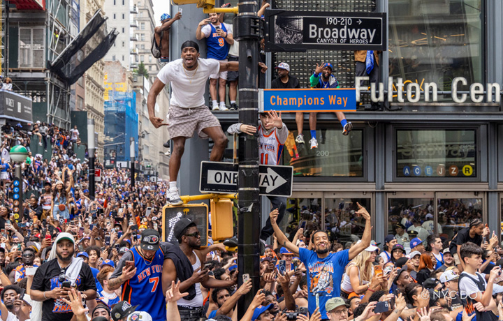 Crowd wearing Knick's gear at Fulton Center with Champions Way and Broadway signs, revelers climbing on poles.
                                           