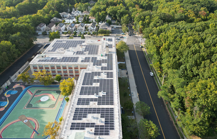 Aerial view of a school building with extensive rooftop solar panels, next to a colorful outdoor playground, surrounded by tree-lined streets and a residential neighborhood.
                                           