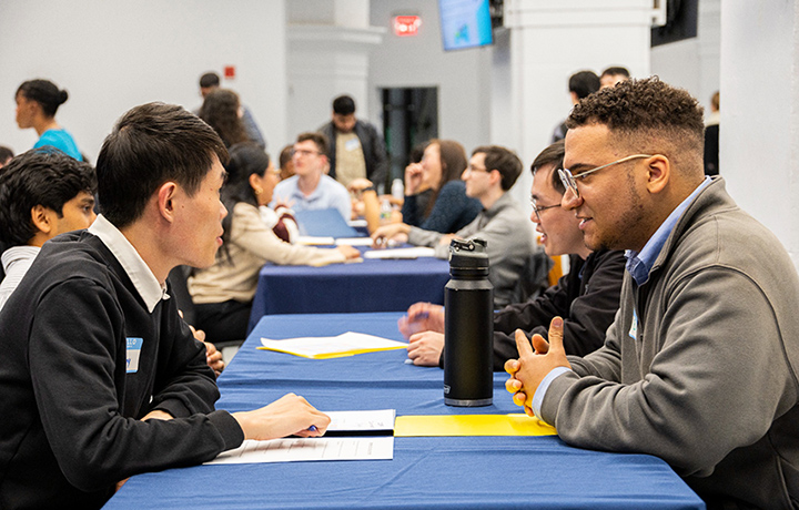 Two DCAS interns sitting across from each other at a blue table, speaking together participating in a mock interview.
                                           