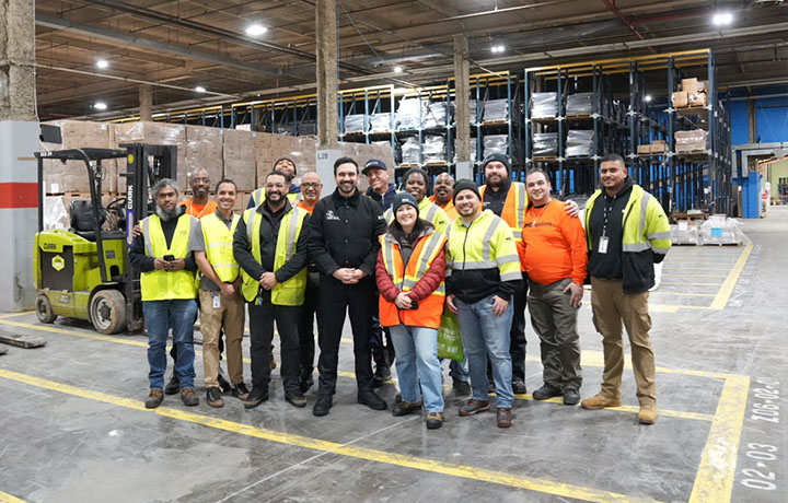 A group of about a dozen workers wearing high-visibility safety vests stand together inside a large warehouse filled with stacked pallets and equipment, posing for a photo near a forklift.
                                           