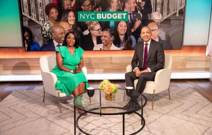 NY1's Cheryl Wills in a green dress and Commissioner Louis Molina in a suit and tie sit together in chairs in front of a glass coffee table. A picture of people and the words NYC Budget is in the background
                                           