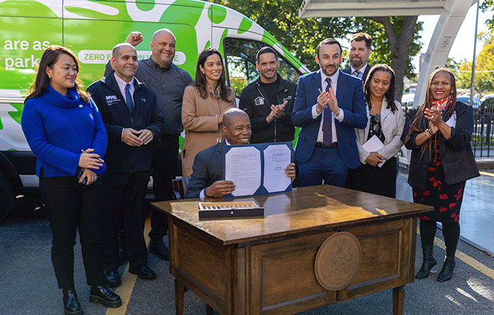 Mayor Adams at desk with signed bill surrounded by senior officials.
                                           