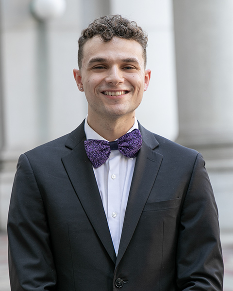 Mitchell is standing outside, smiling, wearing a white shirt, black blazer, and purple bow tie.