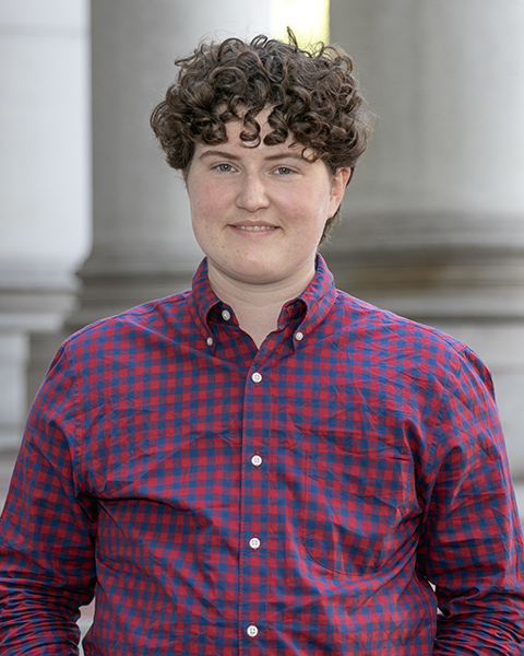 Allison is standing outside, smiling, wearing a blue and red checkered button-down shirt.