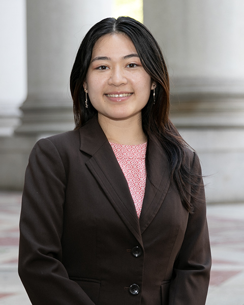 Alysa is standing outside, smiling, and is wearing a pink shirt and brown blazer.