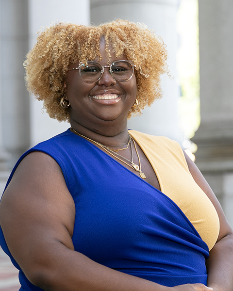 Arielle is standing outside, smiling, wearing glasses and a blue and yellow top.