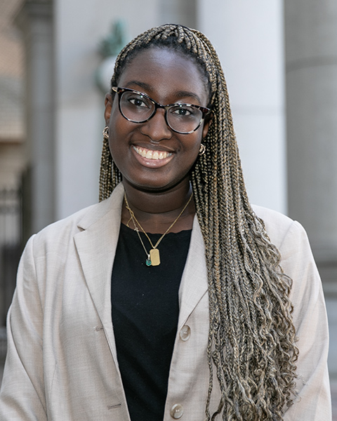 Coura is standing outside, smiling, and is wearing glasses, a black shirt and beige blazer.