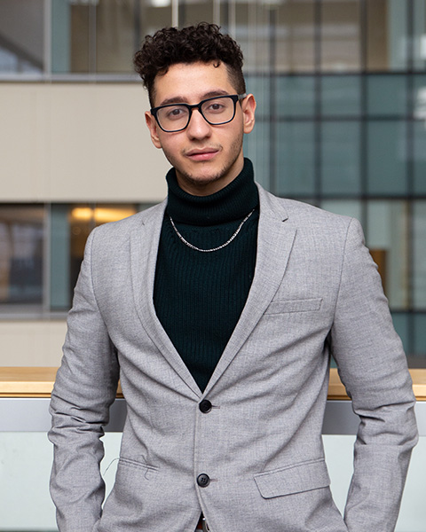 Osvaldo is standing, wearing glasses, a black turtleneck shirt, and light grey blazer. The background has windows.