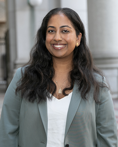 Mehnaz is standing outside, is smiling and wearing a white shirt and light blue blazer.