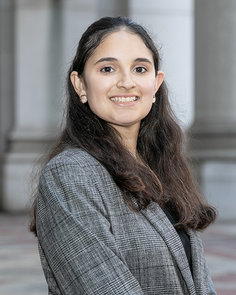 Natalie is standing outside, has  long dark hair is smiling, wearing a grey plaid blazer.