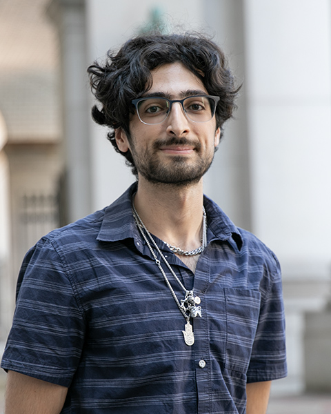 Daniel is smiling, standing outside, wearing silver necklaces and a blue button-down shirt.