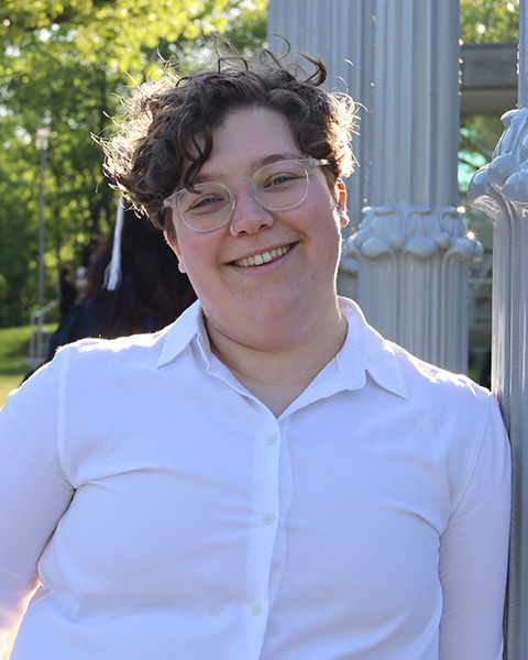 Feigele is standing outside, smiling, wearing glasses and a white button-down shirt.