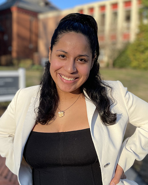 Maria is standing outside, smiling, wearing a black shirt and white blazer.