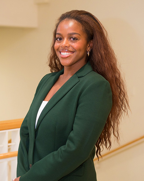 Alanna is standing, smiling, wearing a white shirt and green blazer.