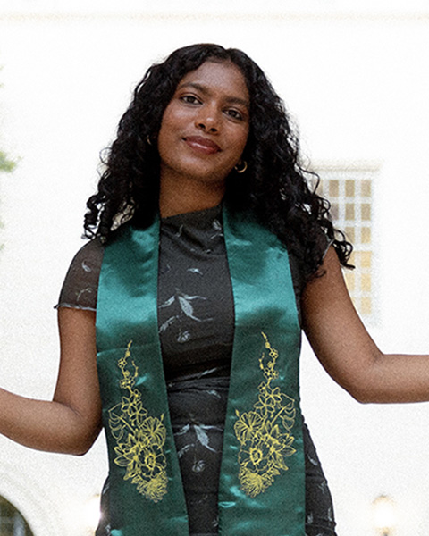 Pritika is standing outside, smiling, wearing a black dress and green sash.