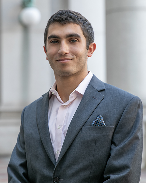 Elliot is standing outside, smiling, wearing a pink shirt with a grey blazer.